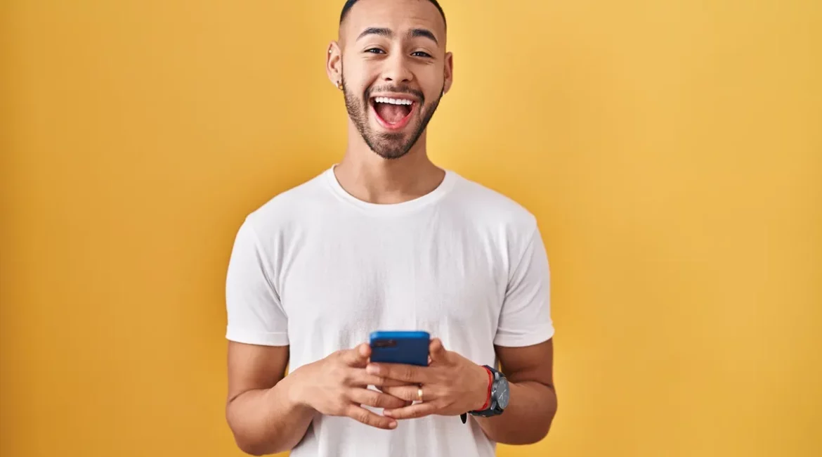 Homem sorridente com barba e camiseta branca segurando um celular azul, em pé contra fundo amarelo, transmitindo alegria e entusiasmo.