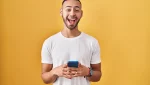 Homem sorridente com barba e camiseta branca segurando um celular azul, em pé contra fundo amarelo, transmitindo alegria e entusiasmo.