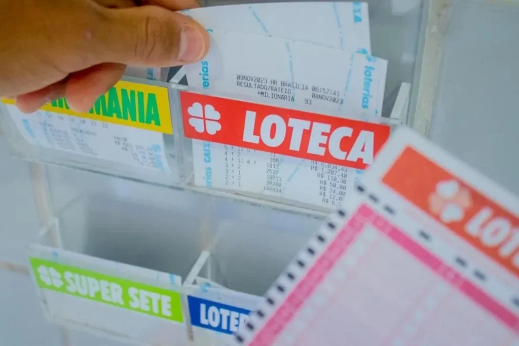 Mão retirando comprovante da Loteca em caixa de acrílico de lotérica com volantes e resultados visíveis, ao lado de bilhetes da Lotomania e Super Sete.
