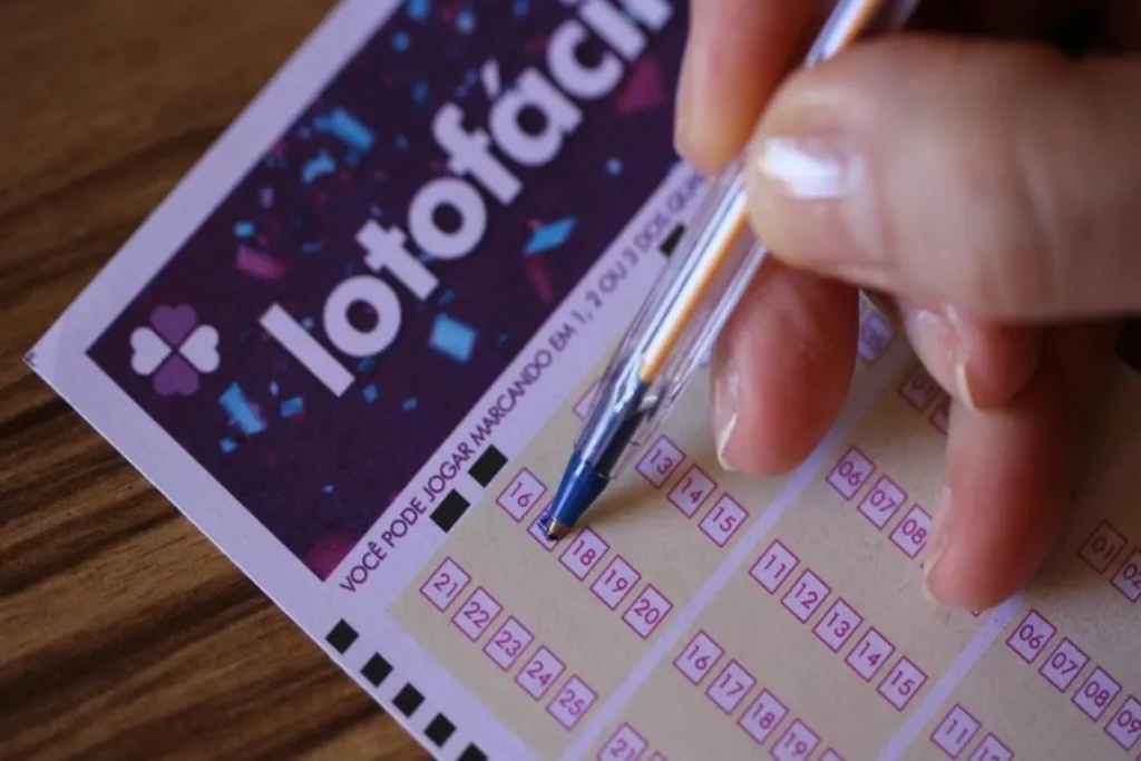 Mão preenchendo um bilhete da Lotofácil com caneta esferográfica azul, simbolizando apostas e jogos de loteria.