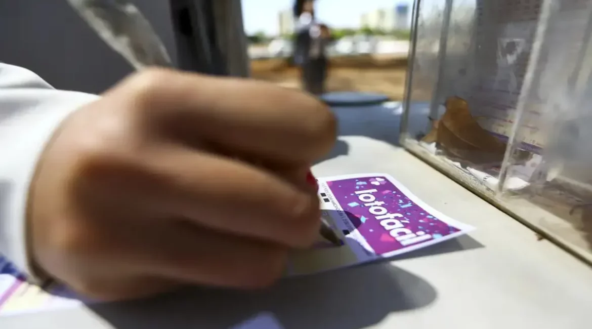 Pessoa preenchendo manualmente um volante da Lotofácil em uma mesa ao ar livre, com urna de apostas ao lado