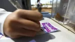 Pessoa preenchendo manualmente um volante da Lotofácil em uma mesa ao ar livre, com urna de apostas ao lado