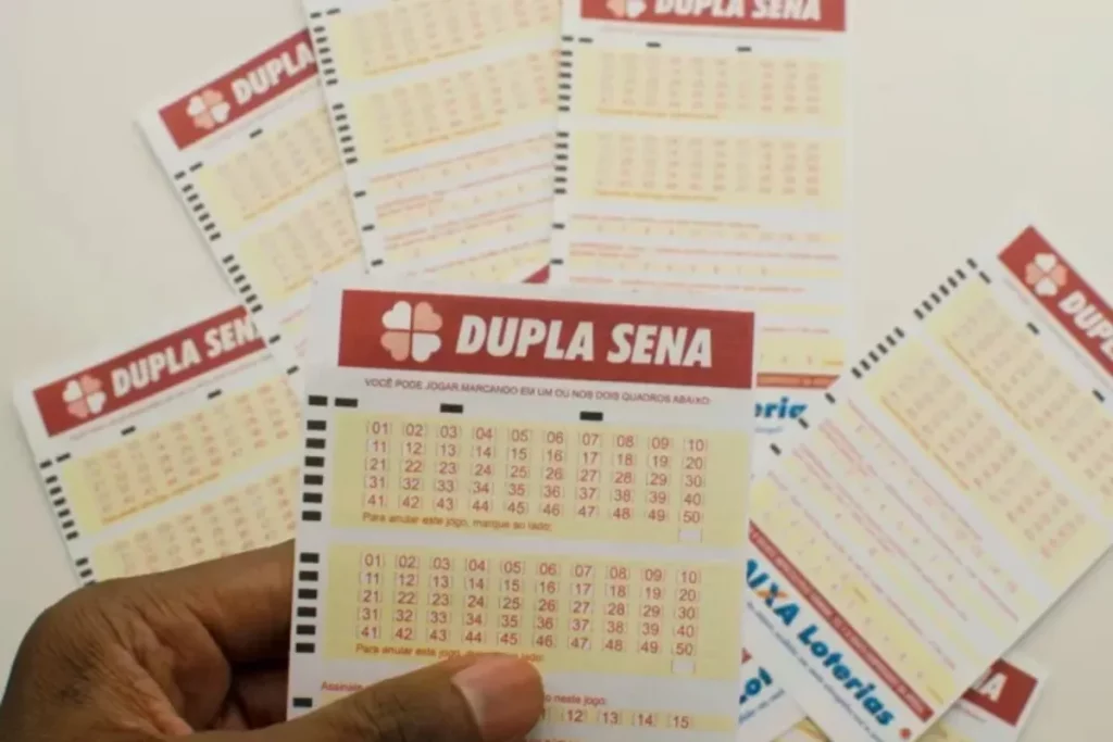 Pessoa segurando volante da Dupla Sena da Caixa Econômica Federal, com vários bilhetes de loteria ao fundo
