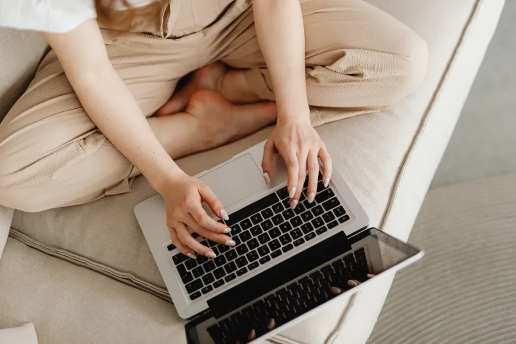 Pessoa sentada no sofá utilizando um laptop no colo enquanto digita no teclado.