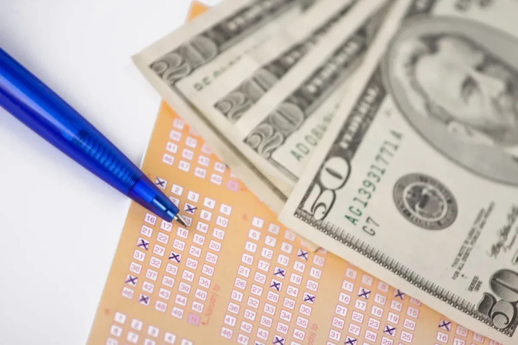 volante de loteria com números marcados ao lado de cédulas de dinheiro e caneta azul